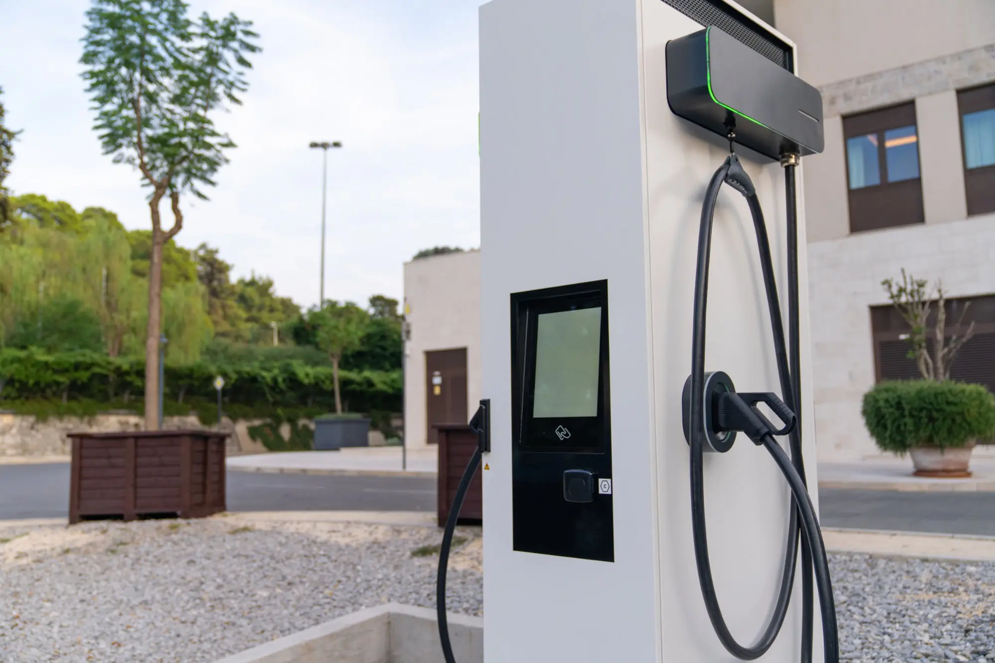 Borne de recharge électrique moderne (EV) blanche avec écran tactile, câbles noirs et lumière verte, sur un parking en gravier devant un bâtiment.